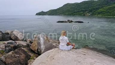 空中盘旋的镜头，金发女孩坐在花岗岩岩石上的瑜伽姿势，周围是异国情调的热带泻湖。